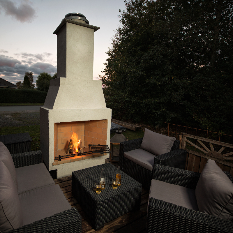 Ochre Living Perisher Complete Fireplace Kit on a timber deck at dusk, with outdoor wicker lounge seating and a lit fire creating a cosy entertaining space