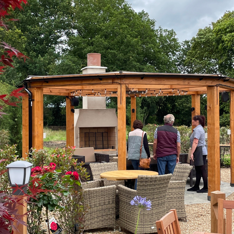 Ochre Living Perisher Complete Fireplace Kit displayed in an outdoor garden showroom beneath a timber pergola, surrounded by outdoor furniture and visitors