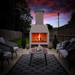 Evening lifestyle image of the Ochre Living Perisher Complete Fireplace Kit with a lit fire, shown in a backyard entertaining area under a dramatic sunset sky