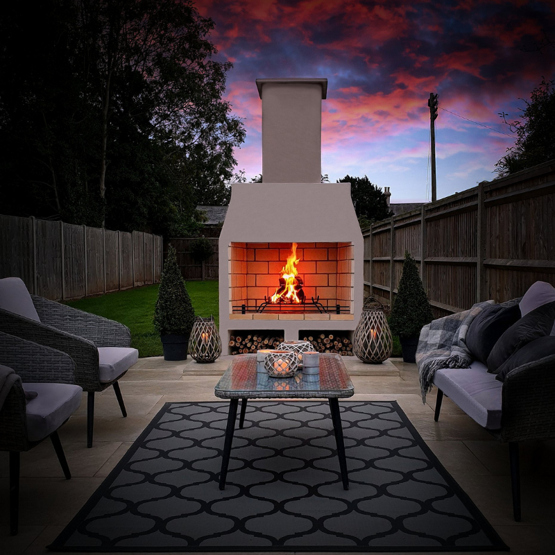 Evening lifestyle image of the Ochre Living Perisher Complete Fireplace Kit with a lit fire, shown in a backyard entertaining area under a dramatic sunset sky