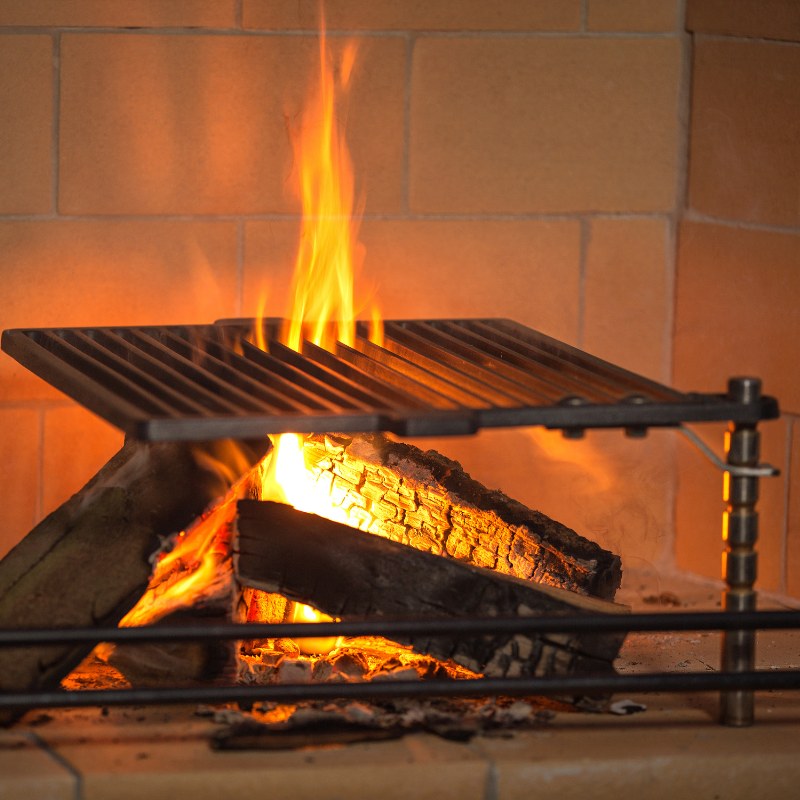 Close-up of the Ochre Living Perisher Complete Fireplace Kit firebox with a grill grate above burning logs, highlighting its wood fire cooking and heating functionality.