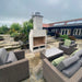 Modern courtyard installation of the Ochre Living Perisher Complete Fireplace Kit, surrounded by outdoor wicker seating on a paved terrace in a landscaped backyard.