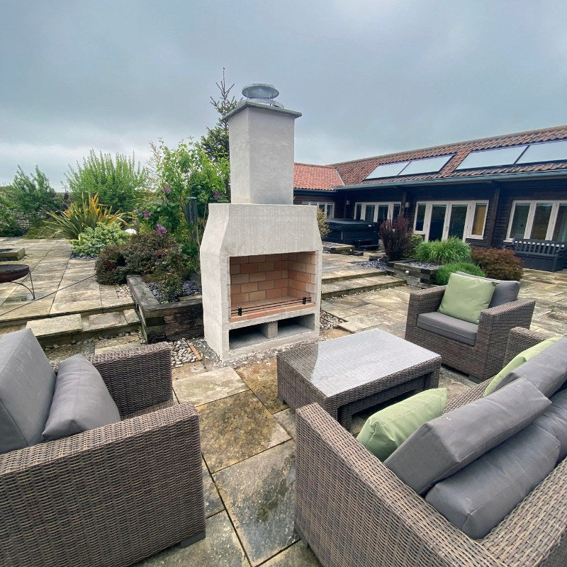 Modern courtyard installation of the Ochre Living Perisher Complete Fireplace Kit, surrounded by outdoor wicker seating on a paved terrace in a landscaped backyard.