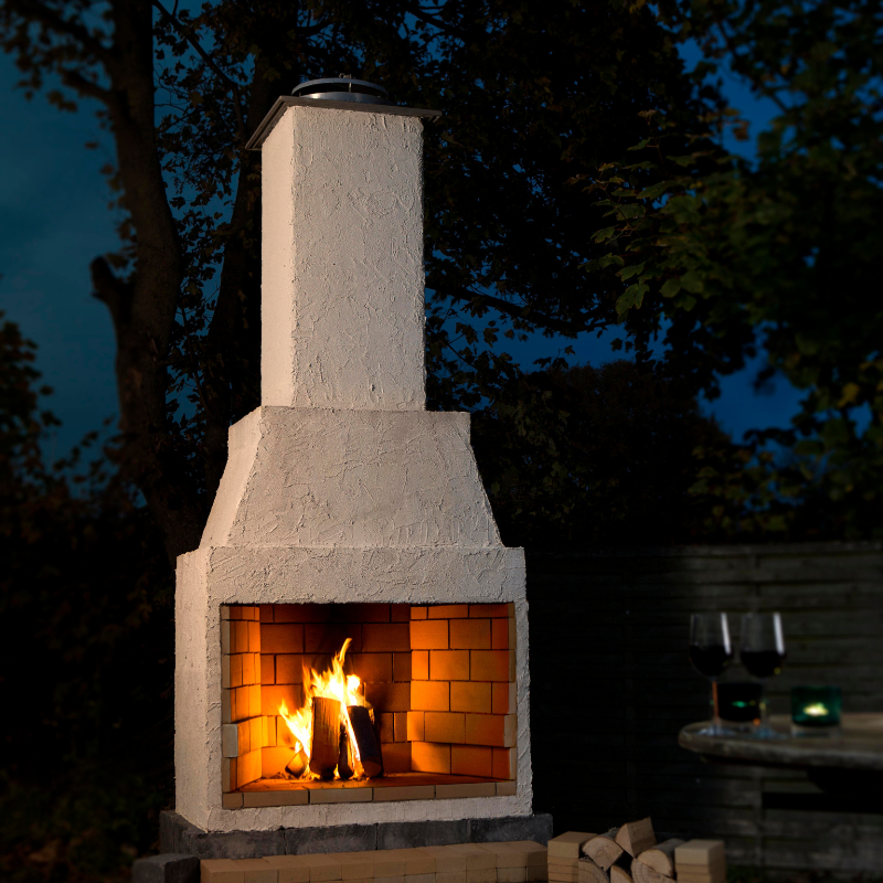 Night-time close view of the Ochre Living Perisher Complete Fireplace Kit with a glowing wood fire, textured rendered surround, and stacked firewood nearby.