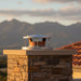Ochre Living Rain Cap installed on a stone chimney outdoors with mountain landscape in the background, showing how the chimney cap protects against rain while maintaining airflow