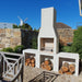 Garden view of the Ochre Living Ossa Complete Fireplace Kit installed beside an outdoor dining setting, with log storage and chimney visible in a landscaped Australian backyard.