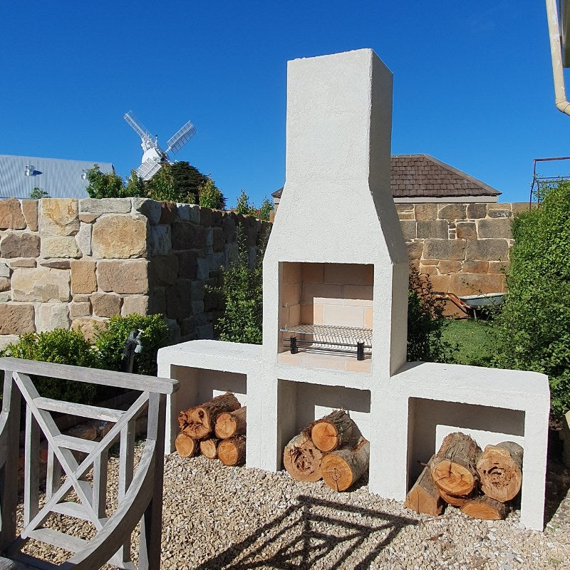 Garden view of the Ochre Living Ossa Complete Fireplace Kit installed beside an outdoor dining setting, with log storage and chimney visible in a landscaped Australian backyard.
