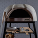 Front detail of the Polito Antonius Garden Wood Fired Oven featuring the stainless steel oven opening, timber pizza peels, pizza thermometer, and preparation shelf on the stand
