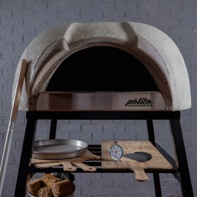 Front detail of the Polito Antonius Garden Wood Fired Oven featuring the stainless steel oven opening, timber pizza peels, pizza thermometer, and preparation shelf on the stand