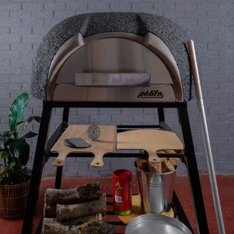 Front view of the charcoal Polito Antonius Garden Wood Fired Oven with stainless steel door, pizza oven tools, timber peels, firewood bucket, and preparation accessories