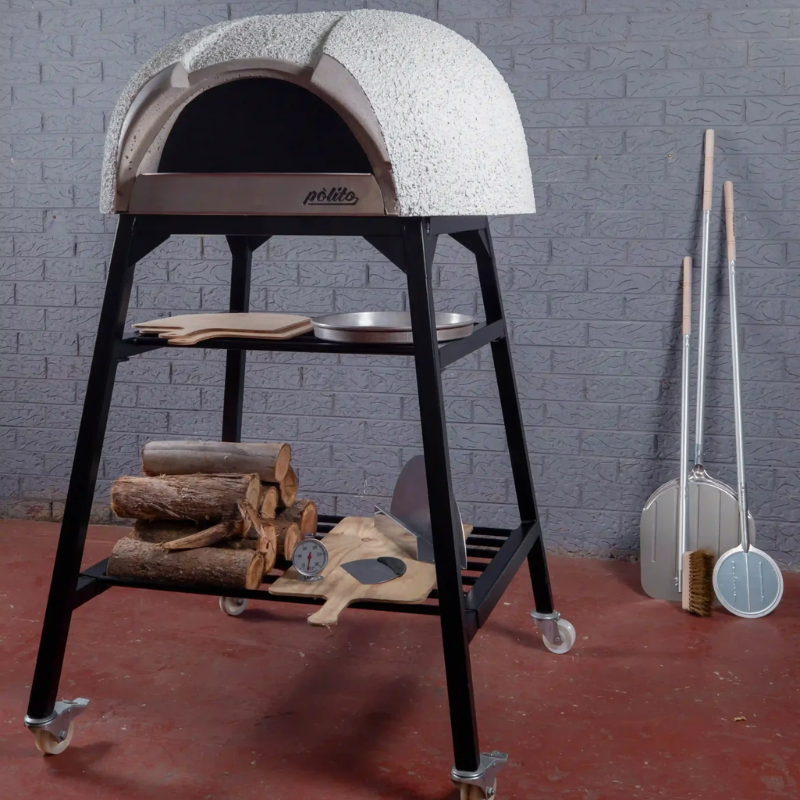 Polito Antonius Garden Wood Fired Oven styled with a Polito pizza oven stand, stacked firewood, pizza thermometer, peels, and stainless steel pizza tools beside the oven
