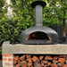 Polito Giotto wood fire oven in black on its screw-in feet, fire lit, on a concrete bench above stacked firewood
