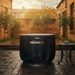 Replacement VisionLINE Fire Pit Cover fitted over a round fire pit in a paved outdoor courtyard, with wet tiles, garden trees, string lights, and a small side table nearby.