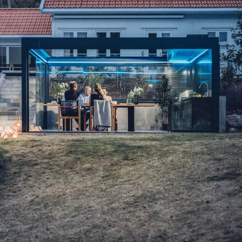 Glass-enclosed outdoor room built with a SkyFlex Delta Open Sky pergola, illuminated with blue LED lighting and used as an evening dining space