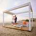 White SkyFlex Delta Open Sky motorised aluminium louvred pergola installed on a beachside platform with lounge seating and two people standing underneath