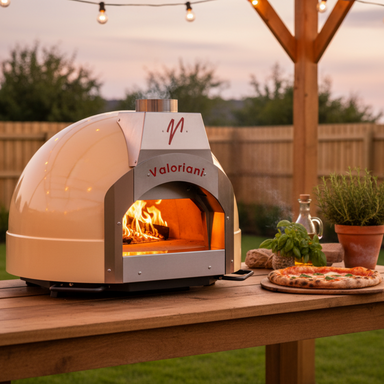 A side-profile view of a cream-colored, built-in Valoriani Family BABY F60 pizza oven for home, actively burning a wood fire. It's positioned on a rustic wooden table at sunset, with a freshly cooked margherita pizza on a paddle next to it, fresh basil, olive oil, and rosemary in a pot. Overhead, string lights glow against a garden background.