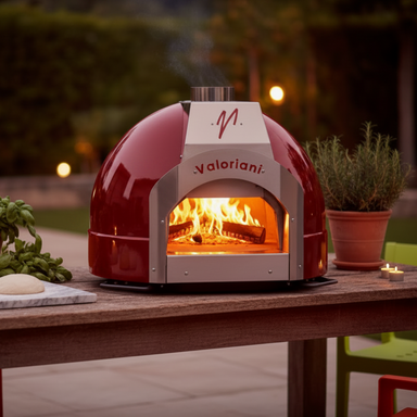 A front-facing shot of a red, built-in Valoriani wood fired oven (specifically the Family BABY F60 Pizza Oven), glowing with internal firelight at dusk. It is set on a wooden patio table, flanked by a pot of rosemary, a plate of dough, fresh basil, and two small lit candles, with green patio chairs and string lights visible.