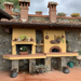 A luxury outdoor kitchen featuring a built-in Valoriani Family FVR120 Pizza Oven integrated into a yellow stucco wall with a stone base, terracotta tile accents, and hanging copper pans.
