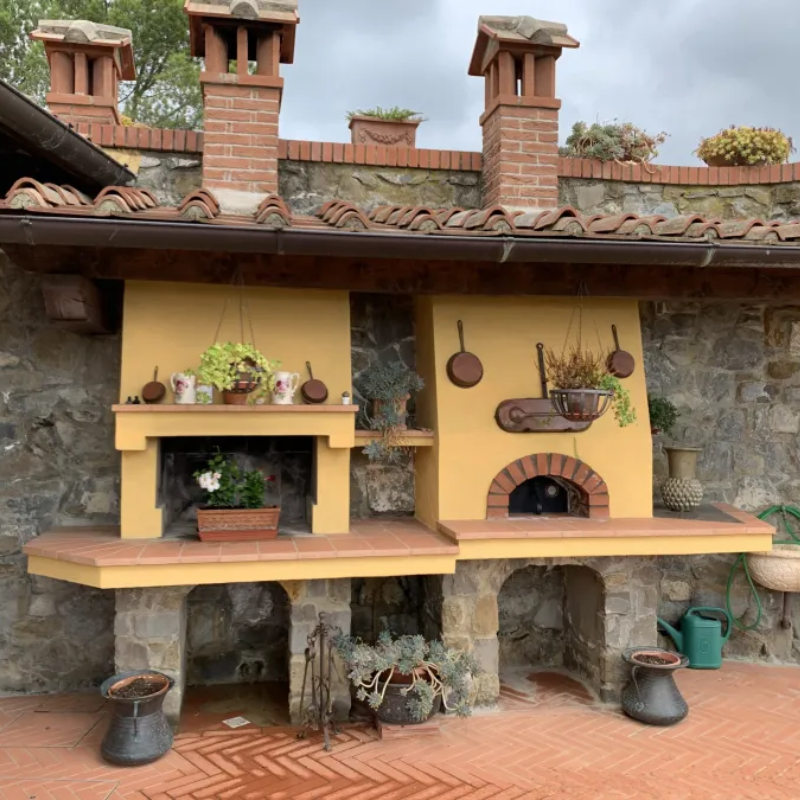 A luxury outdoor kitchen featuring a built-in Valoriani Family FVR120 Pizza Oven integrated into a yellow stucco wall with a stone base, terracotta tile accents, and hanging copper pans.