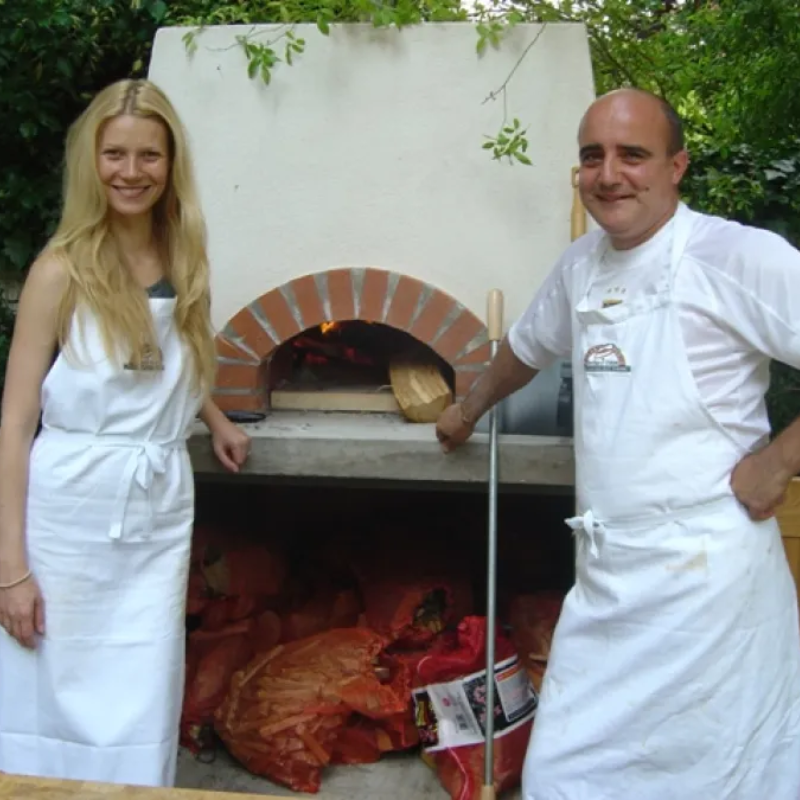 A lifestyle review image showing Gwyneth Paltrow and a chef standing beside a wood-fired Valoriani oven, demonstrating its use in high-end residential outdoor cooking spaces.