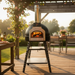 A Black Valoriani Family STAND F75 pizza oven set in a lush outdoor garden during sunset. The oven is placed on a rustic wooden countertop with a freshly baked pizza resting on a wooden board nearby. The scene illustrates the oven as a centerpiece for outdoor dining, available as a Valoriani Family STAND F75 pizza oven under $25,000.