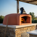 A professional installation of a Valoriani oven in a Mediterranean-style outdoor kitchen—integrated into a stone wall with a terracotta-tiled roof and rustic timber shelving.