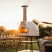 White Valoriani STAND F60 Pizza Oven on a black stand at sunset, with a tall stainless steel flue, warm flame inside, outdoor dining table behind it, and bread on a nearby stone bench.