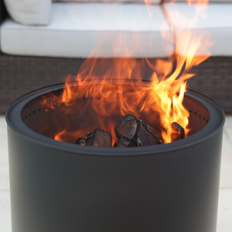 Close-up of the VisionLINE Fire Pit Mini V2 in black, showing orange flames rising above charred wood inside the round fire pit body.