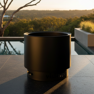 VisionLINE Fire Pit Mini V2 in black positioned on a stone poolside terrace at sunset, with water, native greenery, and a warm outdoor setting behind it.