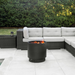 VisionLINE Fire Pit Mini V2 in black burning on a tiled patio, positioned in front of a grey outdoor lounge setting with greenery in the background.