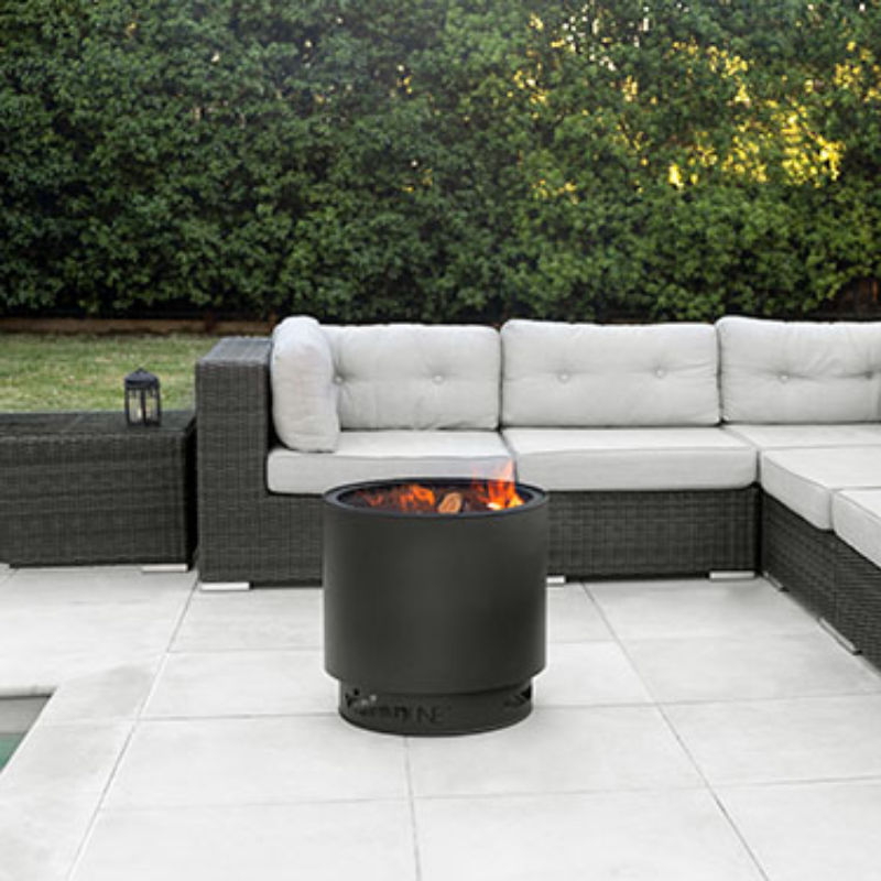 VisionLINE Fire Pit Mini V2 in black burning on a tiled patio, positioned in front of a grey outdoor lounge setting with greenery in the background.