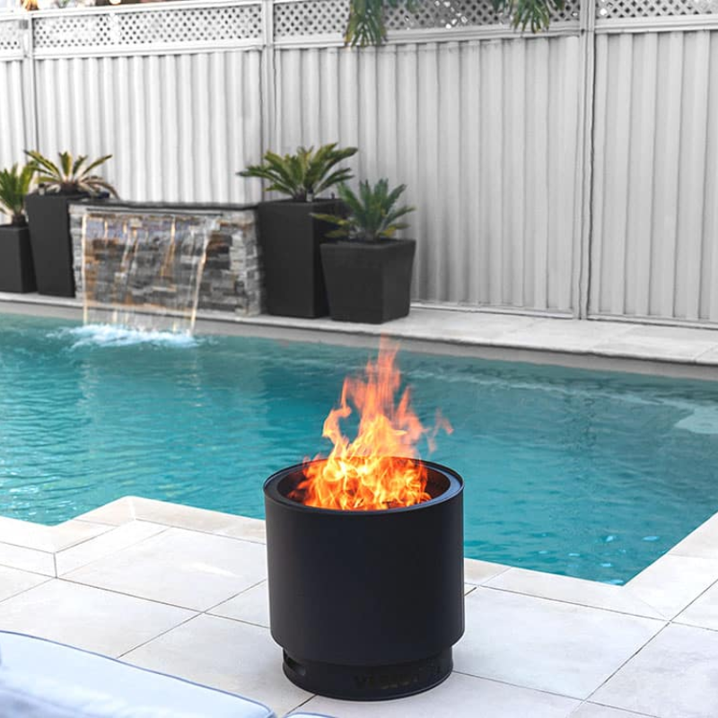 VisionLINE Fire Pit Mini V2 in black burning beside a swimming pool, with bright flames rising from the round fire pit near white outdoor tiles and garden planters.
