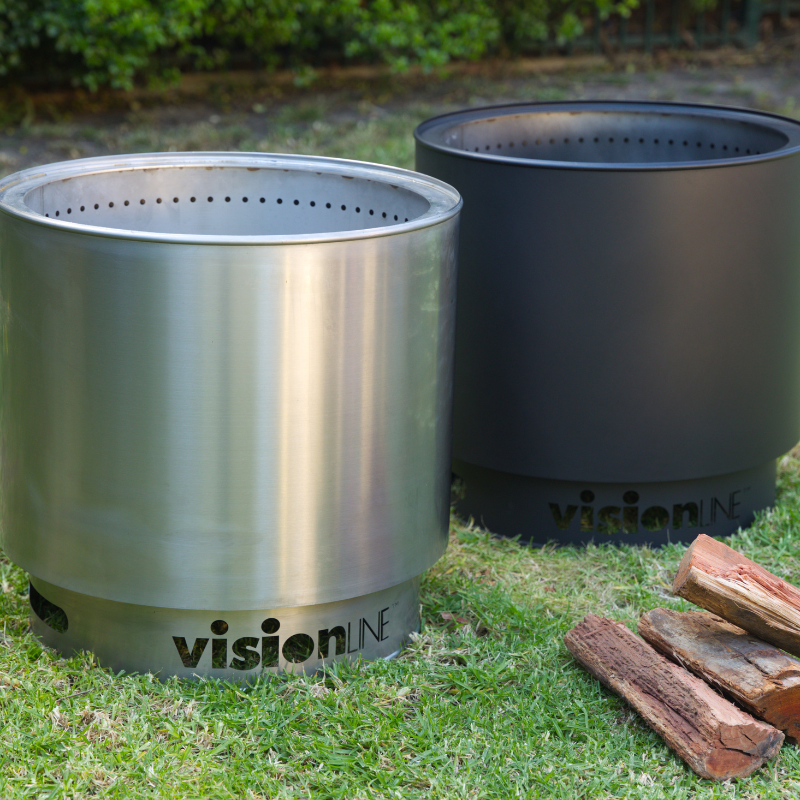 VisionLINE Fire Pit Mini V2 shown in stainless steel and black colour options side by side on grass, with firewood stacked nearby.
