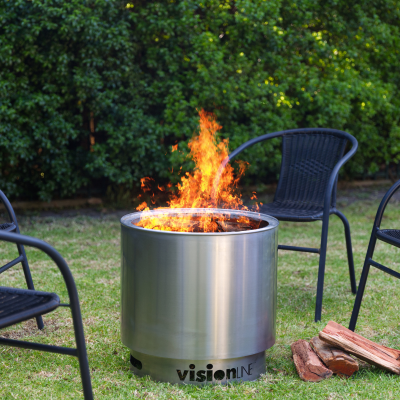 VisionLINE Fire Pit Mini V2 in stainless steel burning on grass, placed between black outdoor chairs with stacked firewood beside the fire pit.