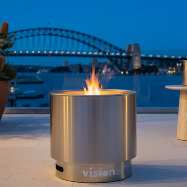 VisionLINE Fire Pit Mini V2 in stainless steel burning on an outdoor balcony at dusk, with a harbour bridge and city lights softly blurred in the background.
