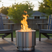 The VisionLINE Fire Pit V2 made of stainless steel, glowing with fire in a backyard setting, surrounded by outdoor seating and illuminated by string lights.