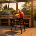 A Wood Valoriani Family STAND F75 pizza oven in a lifestyle setting at dusk. The red oven is positioned on a wooden patio table, glowing from the wood fire within. Surrounding the oven are fresh ingredients like basil, rosemary, and pizza dough, creating an atmosphere often praised in a Valoriani Family STAND F75 pizza oven review.